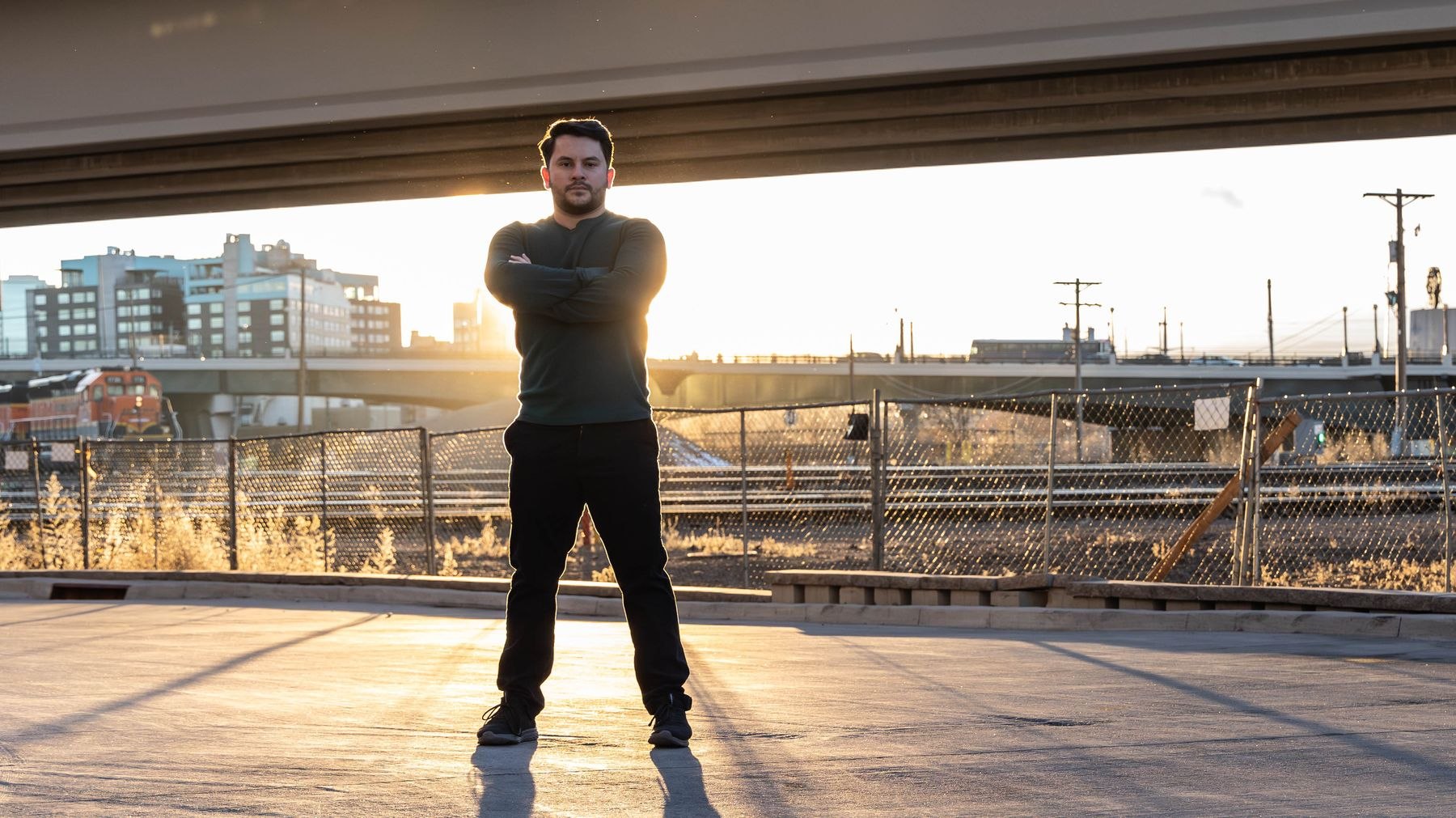 AJ Cassarella at golden hour beneath a Denver overpass, rail lines and skyline behind